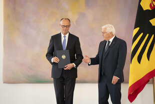 Friedrich Merz steht mit seiner Ernennungsurkunde neben Frank-Walter Steimeier - eine Flagge mit dem Bundesadler ragt ins Bild