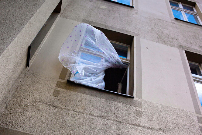 Eine transparente Gardine mit lila Tupfen weht aus dem Fenster eines Mehrfamilienhauses