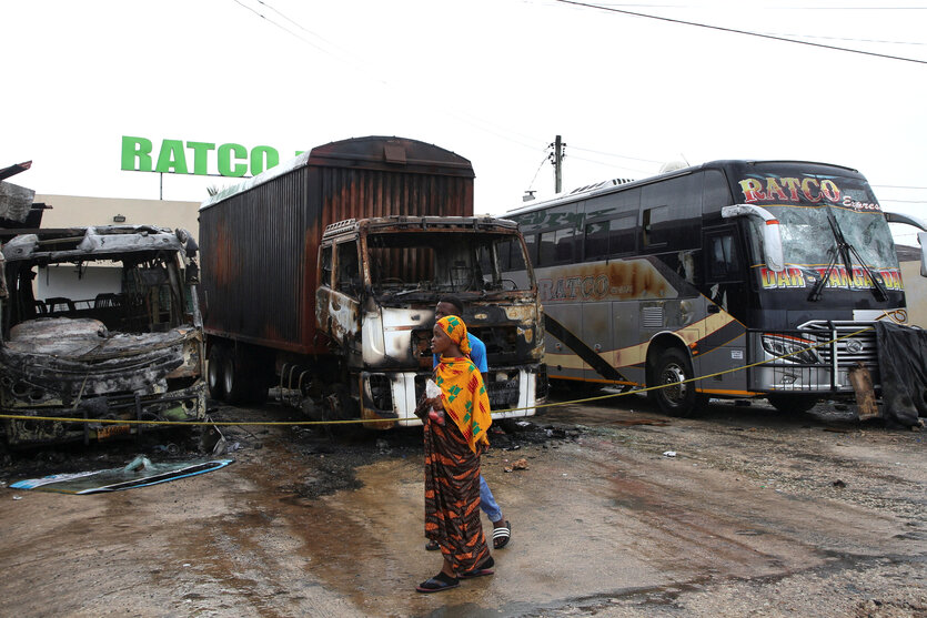 Eine junge Frau in bunter Kleidung geht an einer Reihe von ausgebrannten Bussen vorbei