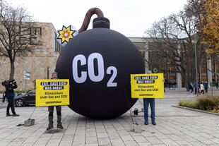 Zwei Personen mit Protestschildern stehen vor einer überdemesional großen Bombenattrappe, auf der "CO2" steht