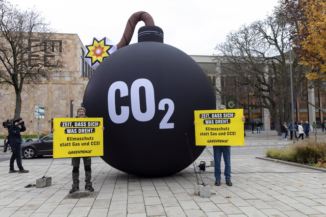 Zwei Personen mit Protestschildern stehen vor einer überdemesional großen Bombenattrappe, auf der 