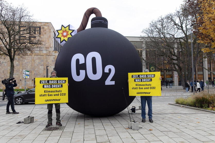 Zwei Personen mit Protestschildern stehen vor einer überdemesional großen Bombenattrappe, auf der "CO2" steht