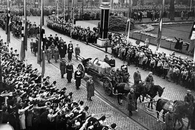 Eine Pferdekutsche transportiert einen Sarg, uniformierte Nationalsozialisten stehen Spalier und zeigen den Hitlergruß