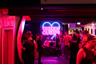 Menschen in rotem Licht im Club, an der Wand grüßt die Leuchtschrift "Cheers Queers"