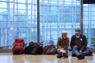 Zwei Menschen sitzen an eine Glaswand angelehnt auf dem Boden eines Flughafenrterminals, neben ihnen reiht sich Gepäck auf
