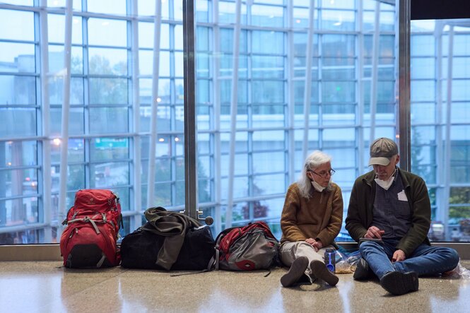 Zwei Menschen sitzen an eine Glaswand angelehnt auf dem Boden eines Flughafenrterminals, neben ihnen reiht sich Gepäck auf