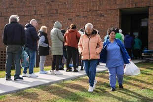Eine Menschenschlange steht vor einem Gebäude, das ein älteres Paar verlässt. Die Frau trägt eine Plastiktüte mit Lebensmitteln