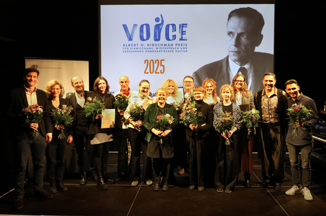 Eine Gruppe von Menschen mit Blumen, im Hintergrund ein Foto von Albert O. Hirschman