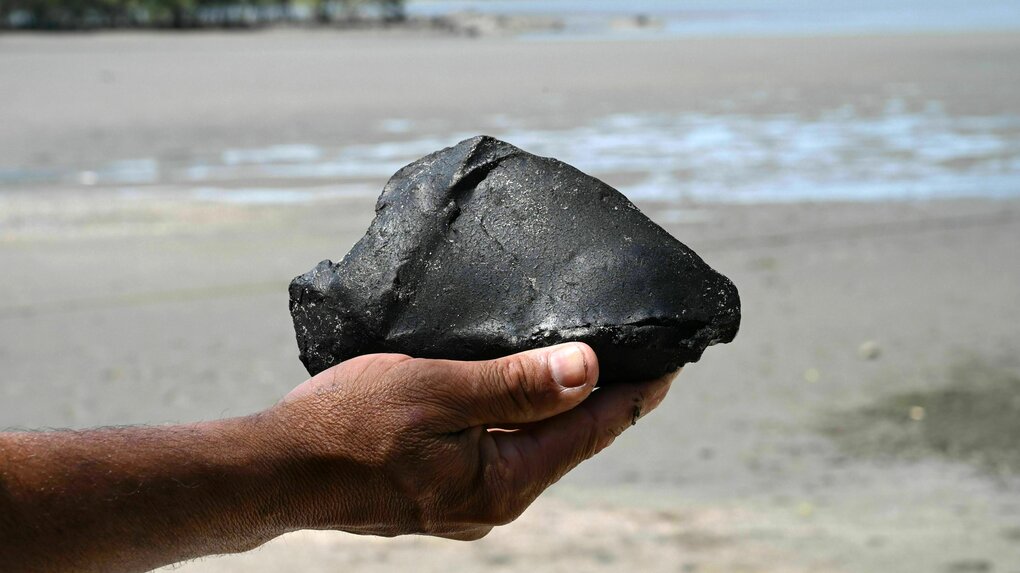 Eine männliche Hand hält einen großen Klumpen Öl in der Hand, im Hintergrund ist ein Strand zu erkennen