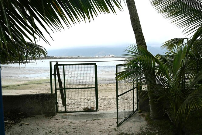 Strandzugang - ein halbgeöffnetes Gitter, dahinter der Strand und Blick auf den Hafen, im Vordergrund Palmwedel