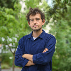 Tom Dubois ist eine weiße Person mit kurzen braunen und wuscheligen Haaren, braunen Augen und braunem Dreitagebart. Er trägt ein dunkelblaues Jeanshemd, schaut mit leichtem Lächeln in die Kamera und hat die Arme vor der Brust verschränkt. Hinter ihm stehen Bäume mit saftig grünem Laub.