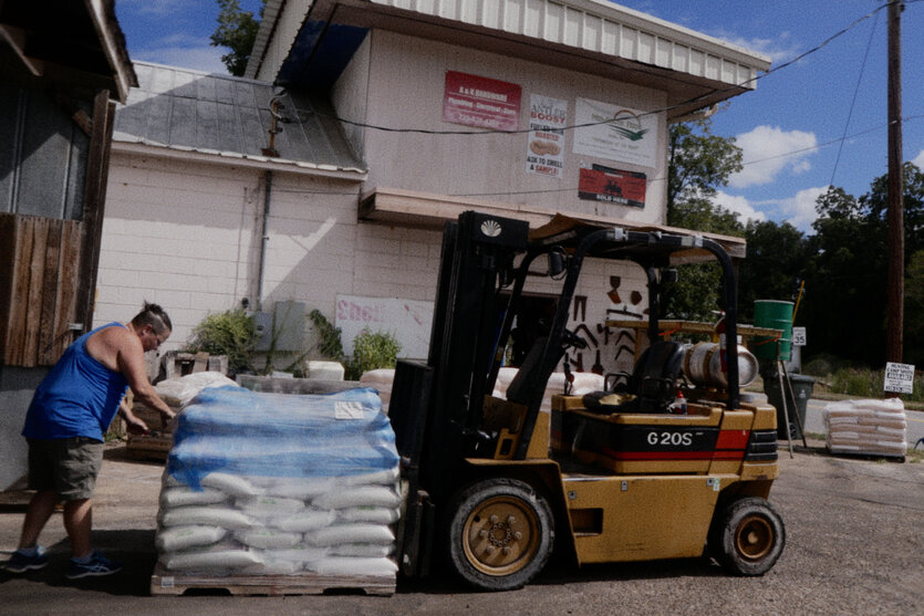 Ein Mann in blauem Tanktop lädt blaue Säcke auf eine Palette vor dem A&K Hardware Store in Lumpkin, Georgia. Ein gelber Gabelstapler mit „G20S“ steht daneben, im Hintergrund Schilder wie „Antler Boost“ und „Soles Here“. Sonniger Tag mit blauem Himmel.