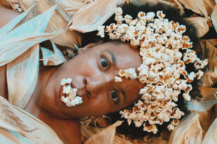 Eine Person liegt in getrockneten Maisblättern, Popcorn auf Haar und Mund. Blick zur Kamera, warmer Ton, surreale Szene.