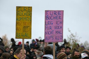 Zwei bunte Pappschilder in einer Menschenmenge: gelb „EURE BÜCHER KOMMEN MIR NICHT INS REGAL“ mit gezeichnetem Buch, rosa „EURE BÜCHER LEG ICH MIR NOCH NICHT MAL AUFS KLO“. Grauer Himmel, winterliche Kleidung.