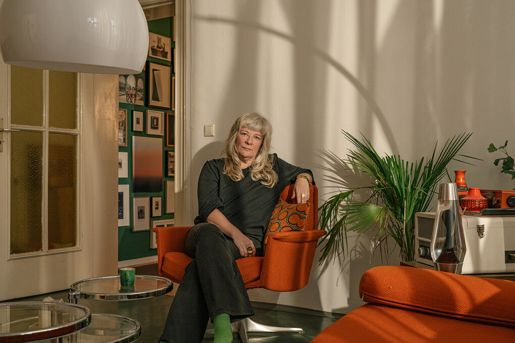 Eine Frau mit grauen haaren sitzt auf einem orangefarbenem Sitzmöbel in ihrer Wohnung