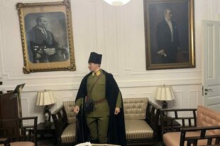 Eine Person in historischer Militäruniform mit Umhang und Mütze steht in einem elegant eingerichteten Raum zwischen zwei großen Porträts an der Wand. Die Einrichtung wirkt klassisch, mit Sesseln, Tischen und Lampen.