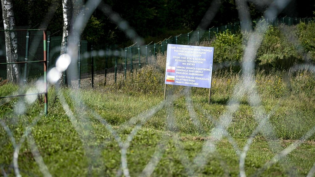 Ein Schild steht an einer Böschung und weist auf die Grenze hin. Im Vordergrund ist Stacheldraht unscharf zu erkennen