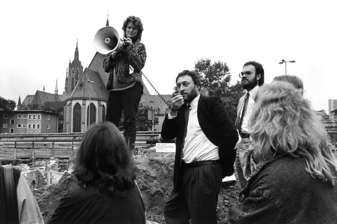 Micha Brumlik mit Jacket und Hemd spricht in ein Megafon, das von einer jungen Frau, die auf einem Erdhügel steht, gehalten wird. Auf dem Bild sind noch vier weitere Personen zu sehen