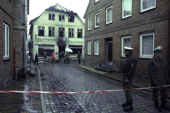 Altes Farbbild; eine verregnete Straße von der Polizei abgesperrt, weitere Rettungskräfte im Hintergrund vor dem abgebrannten Haus