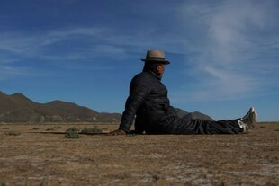Eine Person mit Hut und dunkler Jacke sitzt auf trockenem Boden in einer weiten, kargen Landschaft und blickt zu den fernen Bergen unter einem blauen Himmel.