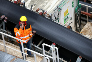 EIn Bauarbeiter in oranger Jacke und gelbem Helm blickt aus einer Baugrube nach oben, dabei in ein Funkgerät sprechend und mit nicht ganz so glücklichem Gesichtsausdruck