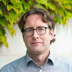 Jakob Graichen ist eine weiße Person mit braun-grauen, kurzen, leicht welligen und gescheitelten Haaren. Er trägt eine schwarze, viereckige Brille und ein hellblaues Hemd. Hinter ihm ist eine helle Mauer, an der grüner Wein wächst.