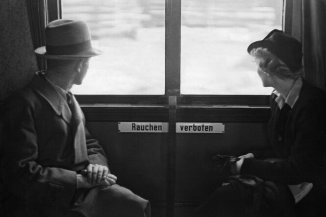 Menschen auf Reisen. Herr und Dame in einem Zugabteil schauen aus dem Fenster (ein "Rauchen verboten'"- Schild zwischen ihnen), in Schwarz-Weiß