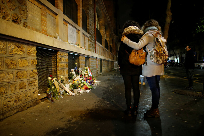 Nacht, zwei weiblich gelesene Personen stehen in Andacht, eine Person hat der anderen den Arm umgelegt, vor niedergelegten Blumen und Kerzen vor einer Mauer mit Fenstern