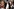Drei Personen in extravaganten 80er-Punk-Outfits und grell geschminkten Gesichtern posieren eng beieinander: Links eine Frau mit wildem Blondhaar, blutigem Make-up und Perücken als BH; in der Mitte ein Mann mit Federn, blauer Gesichtsbemalung und Fellweste, der eine Gitarre hält; rechts ein Mann mit goldbemaltem Gesicht, spitzem Blondhaar und offener Bluse.