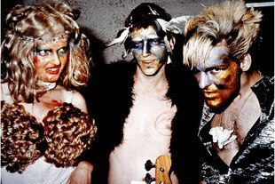 Drei Personen in extravaganten 80er-Punk-Outfits und grell geschminkten Gesichtern posieren eng beieinander: Links eine Frau mit wildem Blondhaar, blutigem Make-up und Perücken als BH; in der Mitte ein Mann mit Federn, blauer Gesichtsbemalung und Fellwest