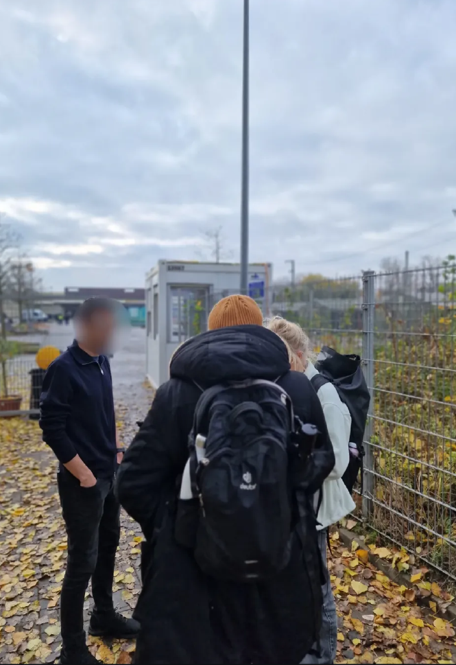 Drei Menschen vor einem Gitterzaun, in Hintergrund ein Wachcontainer