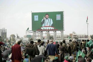 Auf einem Videoscreen ist ein Mann zu sehen, der mit dem Zeigefinger in die Höhe zeigt, es ist Abdulmalik al-Huthi