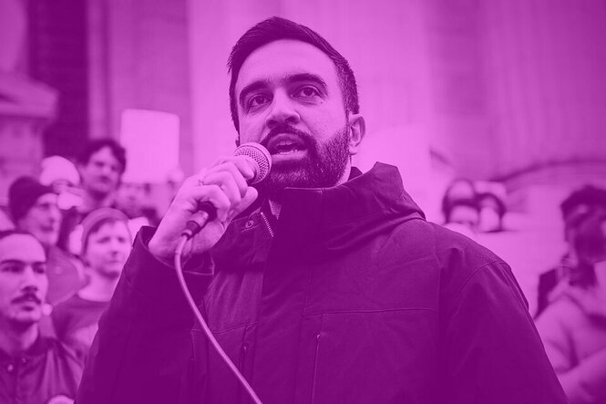 Zohran Mamdani spricht mit einem Mikrofon bei einer Kundgebung vor einer Gruppe von Menschen, die teilweise Schilder mit auf dem Foto unkenntlicher Schrift hochhalten. Er trägt einen Mehrtagesbart und hat kurzes, schwarzes Haar.