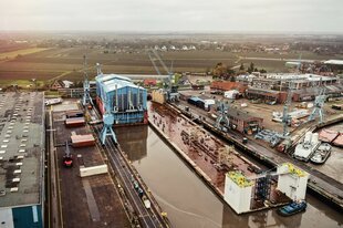 Luftaufnahme einer Werft: Blaue Schiffshalle in einem Hafenbecken, umgeben von Kränen, Containern und Gebäuden.
