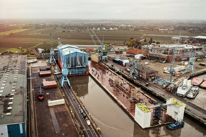 Luftaufnahme einer Werft: Blaue Schiffshalle in einem Hafenbecken, umgeben von Kränen, Containern und Gebäuden.