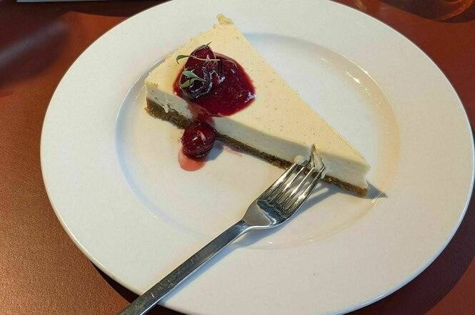 Ein Stück Cheesecake mit roter Fruchtsauce liegt auf einem weissen Porzellanteller, den eine Gabel ziert