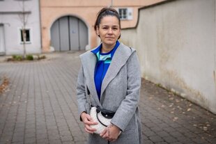 Portraitfoto von Meryem: Die 36-Jährige hat ihre schwarzen Haare zum Zopf gebunden, trägt einen grauen Mantel über einem blauen Oberteil, ihre Hände liegen auf ihrer weiß-beige gemusterten Handtasche.