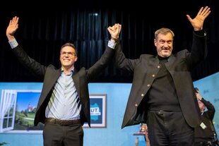 Links steht Manuel Hagel, rechts Markus Söder. Hand in Hand strecken sie auf der Bühne mit türkisblauer Kulisse ihre Arme in die Höhe.