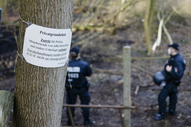 An einem Schild an einem Baum steht: "Privatgrundstück – Zutritt für Polizei, Mitarbeitende von JaRa-Immobilien und andere Unbefugte verboten! Zuwiderhandlungen werden gemäß §123 ff. StGB zur Anzeige gebracht." Im Hintergrund stehen Polizisten im Wald.