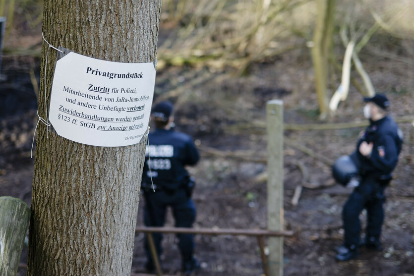 An einem Schild an einem Baum steht: "Privatgrundstück – Zutritt für Polizei, Mitarbeitende von JaRa-Immobilien und andere Unbefugte verboten! Zuwiderhandlungen werden gemäß §123 ff. StGB zur Anzeige gebracht." Im Hintergrund stehen Polizisten im Wald.
