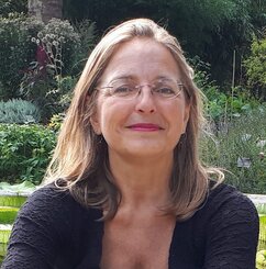 Letizia Paoli. Eine Frau mit blonden Haaren, die ihr bis über die Schultern hängen. Sie trägt eine Brille und ein dunkles Oberteil. Sie lächelt. Im Hintergrund grüne Sträucher und grüner Boden.