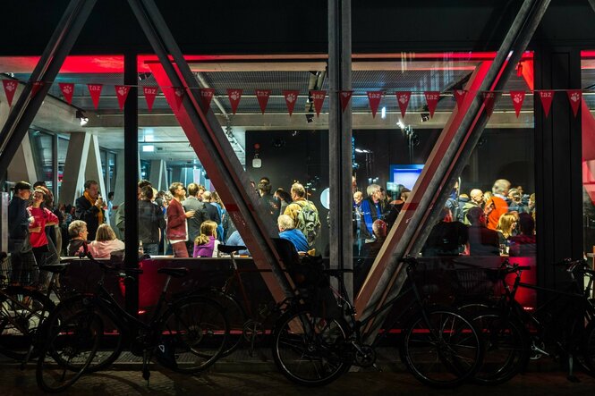 Blick durch eine Glasfassade des taz-Gebäudes: Viele Menschen stehen und sitzen im hell erleuchteten Raum, unterhalten sich und trinken. Vor dem Fenster sind Fahrräder abgestellt, rote Lichter und Wimpel mit taz-Logo schaffen eine lebhafte Atmosphäre.