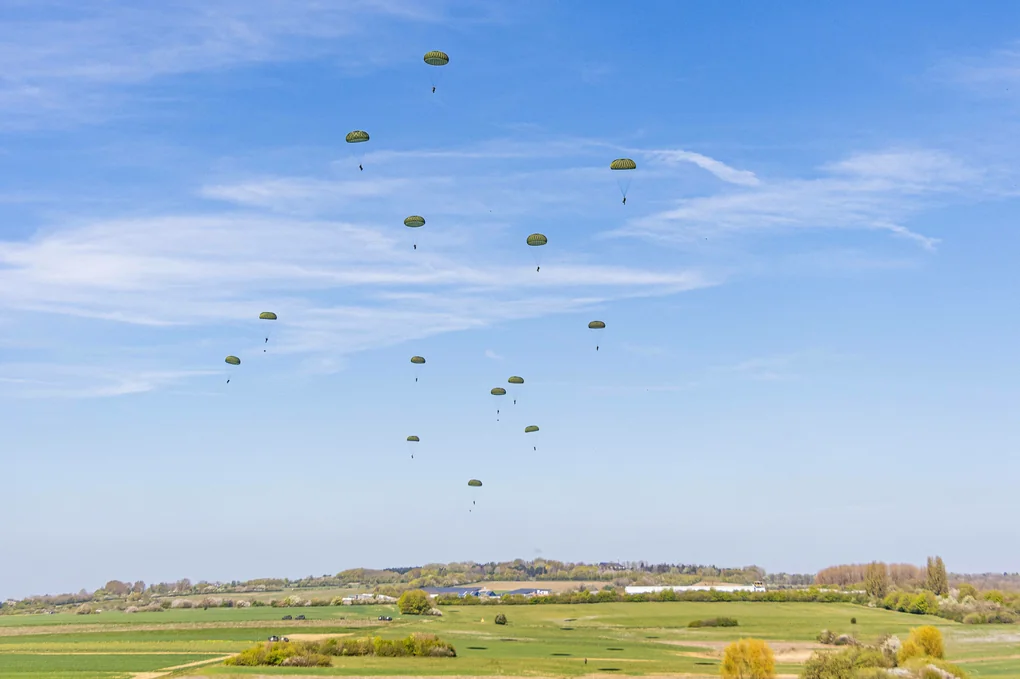 Fallschirmjäger setzten mit ihren Rundkappen auf einer grünen weiten Wiese ab, kontrastiert vom blauen Himmel