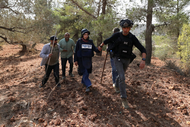 Die verletzte Fotografin wird von einem Kollegen und mehreren Palästinensern begleitet - der Boden ist mit Geroll übersät, Nadelbäume stehen in der Landschaft