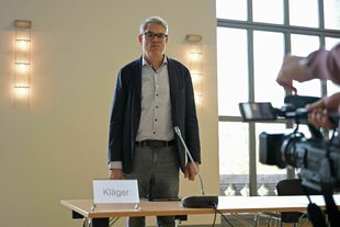 Gerichtssaal, klagenden Seite, ein Herr in schlichtem Sakko und Jeans, mit hellblauen Hemd und dicker Brille