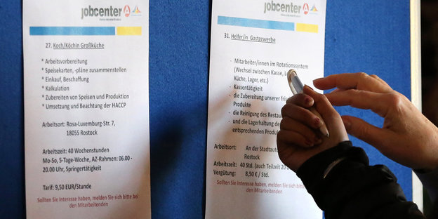 An einem Aufsteller hängen Zettel mit Jobangeboten, zwei Hände zeigen auf einen der Zettel