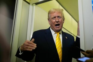 Donald Trump, ein alter Mann reißt den Mund weit auf und hält seine rechte Hand vor sich. Er trägt ein schwarzes Jackette und eine gelbe Jacke. Sein Gesicht ist gelblich. Die Wand hinter ihm ist gelb. Er steht in einem Türrahmen.