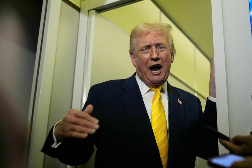 Donald Trump, ein alter Mann reißt den Mund weit auf und hält seine rechte Hand vor sich. Er trägt ein schwarzes Jackette und eine gelbe Jacke. Sein Gesicht ist gelblich. Die Wand hinter ihm ist gelb. Er steht in einem Türrahmen.