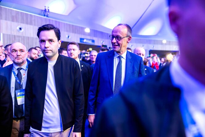 Friedrich Merz geht lächelnd neben Johannes Winkel, der sieht eher schlecht gelaunt aus