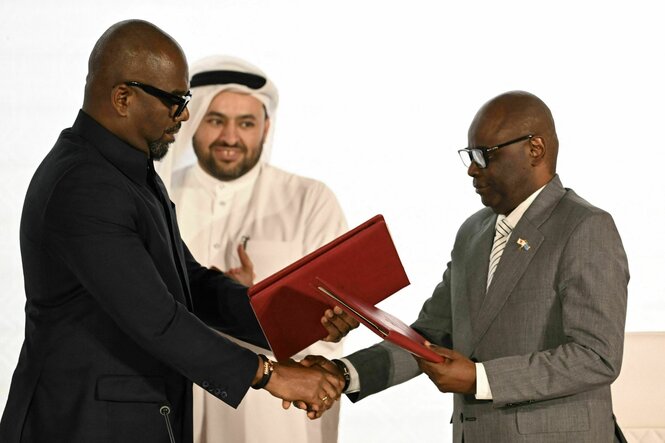 Zwei Männer im Anzug übergeben sich rote Dokumentmappen. Eine Person im Hintergrund klatscht anerkennend